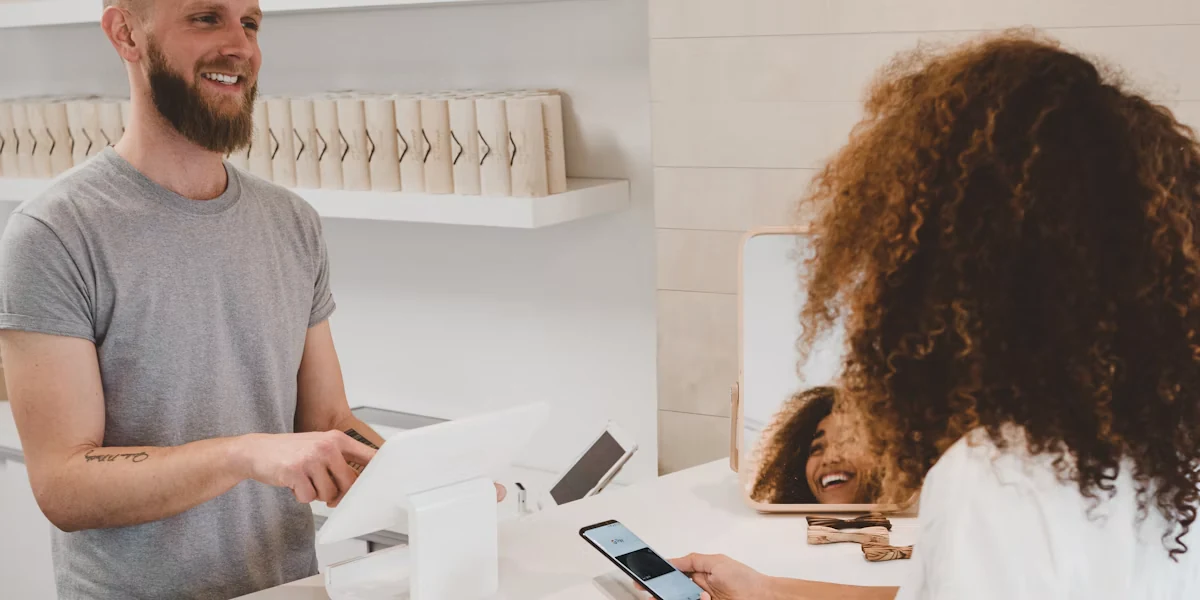 A man uses a white countertop device while a woman holds a smartphone in a bright shop.