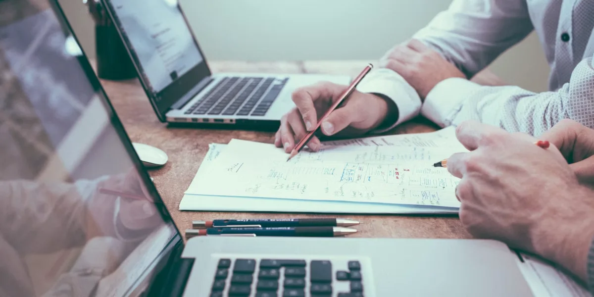 People in business attire write on papers at a wooden table with open laptops in an office.