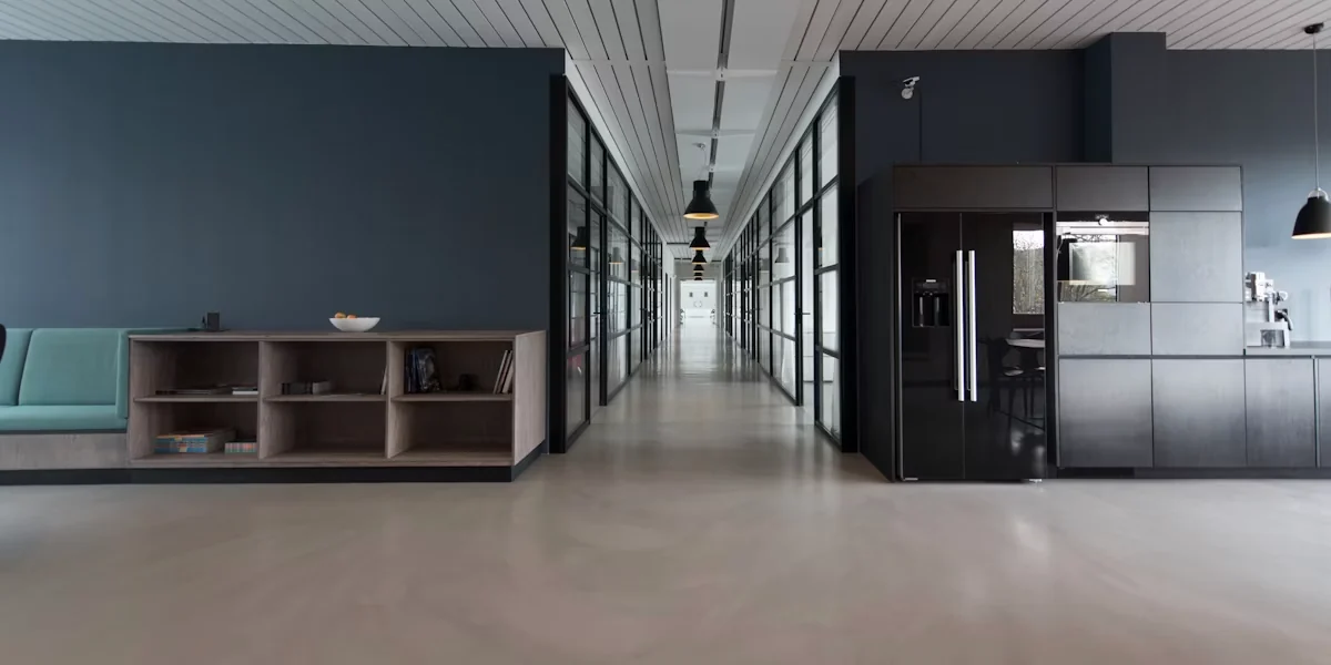Modern commercial office interior with a long glass-walled corridor, dark cabinetry, teal seating, and an empty gray floor; no people.