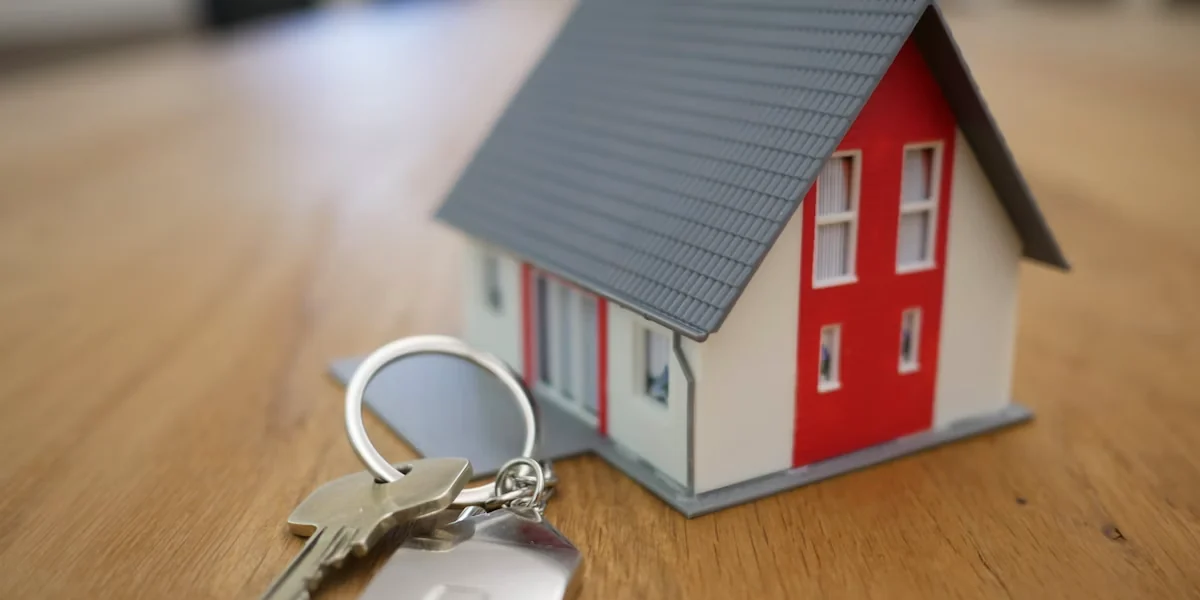 A small model house with red and white walls and a gray shingled roof sits on a wooden surface beside keys.